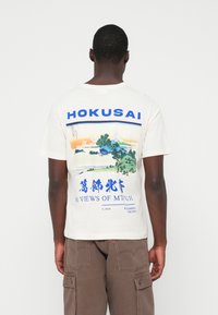 Crème farget bomulls-T-skjorte med et Hokusai-print og blå grafikk og tekst. Den viser en landskapscene med Fuji-fjellet og trær.