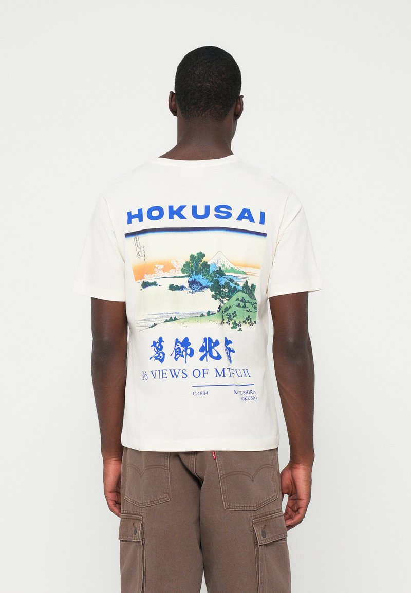 Crème farget bomulls-T-skjorte med et Hokusai-print og blå grafikk og tekst. Den viser en landskapscene med Fuji-fjellet og trær.
