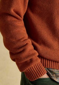 Rostevärviline kudumipusa ribatud varrukaotste ja hemiga, millel on pehme tekstuur. Aluses on ruuduline särgike.