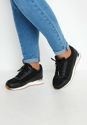 Person trägt blaue hochgekrempelte Jeans und schwarze Schnürsneaker mit weißen Sohlen und braunen Gummiaußensohlen vor weißem Hintergrund.