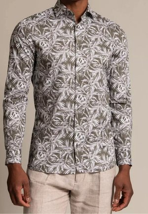 Homme portant une chemise ajustée à manches longues avec imprimé feuilles de palmier blanches sur fond vert olive, associée à un pantalon beige clair.