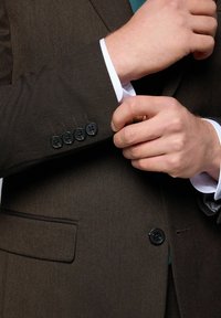 Veste de costume texturée marron avec quatre boutons noirs au poignet, accompagnée d'une chemise blanche en dessous et d'un léger aperçu d'un tissu vert.