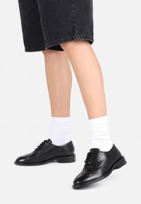 Sorte lærersko med brogue-detaljer, parret med hvite ribbestrikkede sokker, iført mørk denimshorts. Lav hæl-design, jevn tekstur.