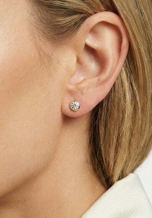 Close-up af en kvindes øre med kort blondt hår, iført en lille rund guldbelagt ørestik med en klar ædelsten.