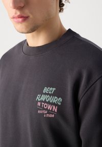 Σκούρος γκρι φούτερ με στρογγυλή λαιμόκοψη. Διαθέτει πολύχρωμο κείμενο "ΚΑΛΥΤΕΡΕΣ ΓΕΥΣΕΙΣ ΣΤΗΝ ΠΟΛΗ" σε πράσινο και ροζ, με την ένδειξη "SCOTCH & SODA" από κάτω.