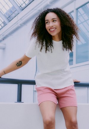 Chemise blanche texturée à manches courtes associée à un short rose clair. Le modèle s'adosse à une rambarde dans un cadre intérieur lumineux.
