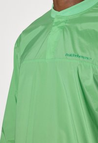 Heldere groene lichte jas met een opstaande kraag en gestikte details. Voorzien van het PeakPerformance-logo in donkergroen op de borst.