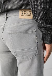 Jeans din denim gri cu o etichetă din piele pe care scrie "SCOTCH & SODA AMSTERDAM." Detalii notabile includ accente cusute pe buzunarul din spate.