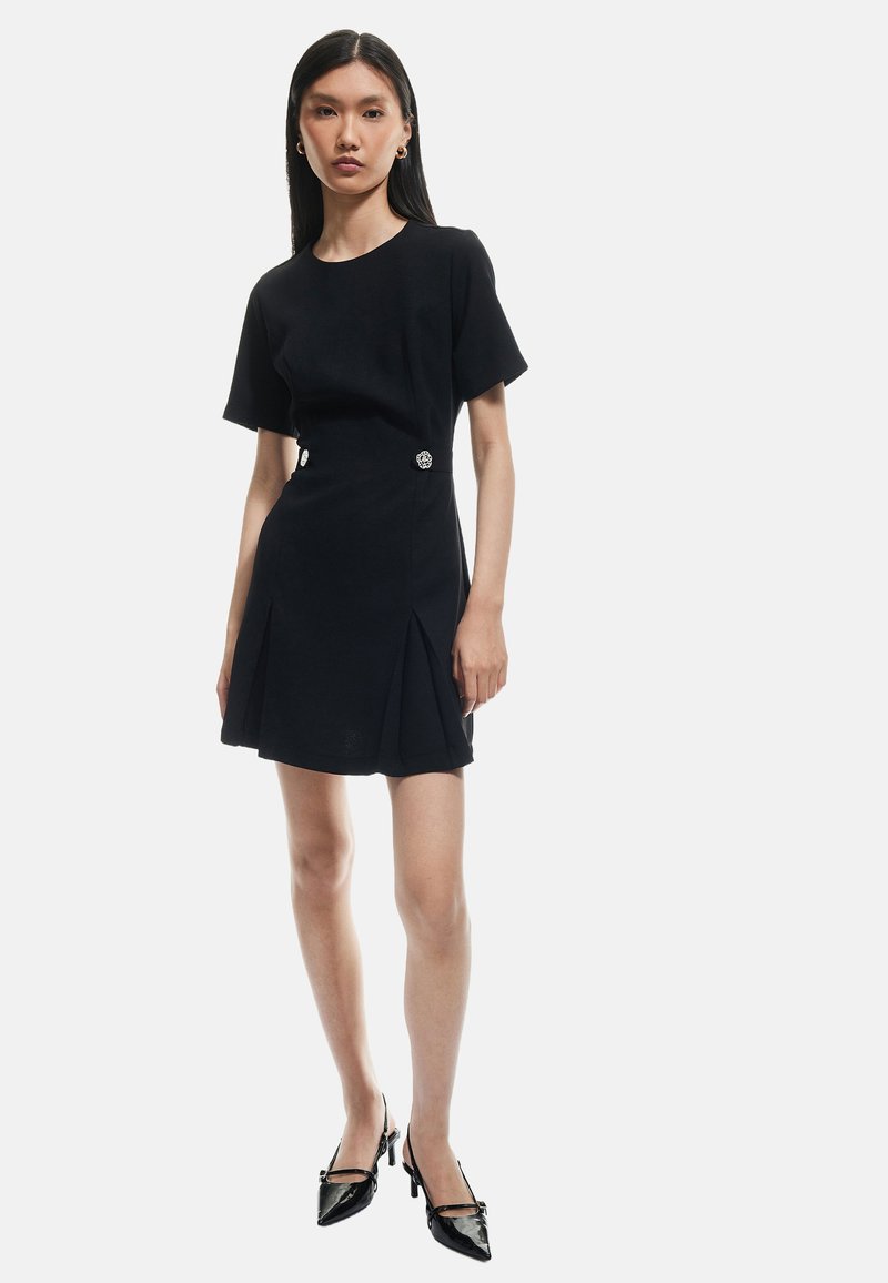 Zwarte jurk met korte mouwen, een getailleerde taille, een geplooide rok en decoratieve knoopdetails aan de zijkant. Gecombineerd met zwarte hakken.
