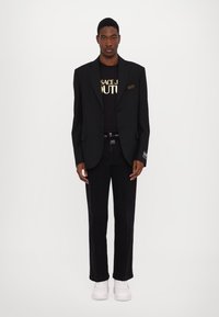 Blazer nero con rever a punta, abbinato a un t-shirt nera con lettering dorato, jeans neri slim fit e sneakers bianche.