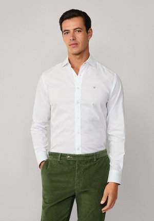 Hombre vistiendo una camisa blanca de botones y pantalones de pana verdes, de pie con una mano en el bolsillo frente a un fondo sencillo.