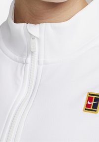 Valkoinen vetoketjullinen kauluspaita, jossa on teksturoidut kangas, etuvetoketju ja pieni monivärinen logo-ompelu.