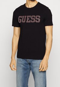 Černé bavlněné tričko s velkým nápisem "GUESS" v červených pruzích. Krátké rukávy, kulatý výstřih a volný střih. Spárováno s modrými džíny.