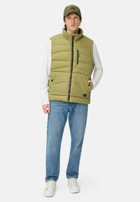Olivgrüne Steppweste mit hohem Kragen, Reißverschlusstasche und Druckknopfverschlüssen, kombiniert mit blauen Jeans und weißen Sneakern, was einen lässigen Look vervollständigt.