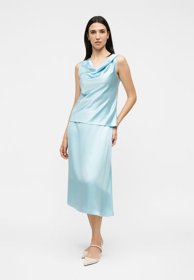 Γυναίκα που φοράει αμάνικη ανοιχτό μπλε σατέν μπλούζα και αντίστοιχη φούστα μήκους midi, στέκεται με το ένα χέρι στην τσέπη της φούστας και άσπρα μυτερά παπούτσια.