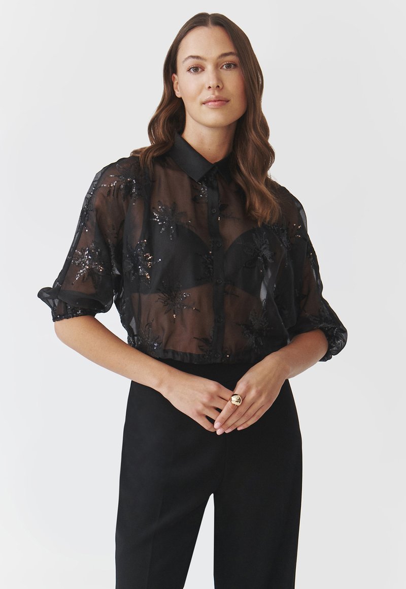 Bluză neagră transparentă cu mâneci scurte, embellisment cu strasuri florale, design cu butoni în față și guler contrastant. Asortată cu pantaloni negri.
