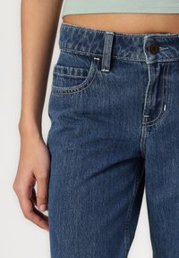 Jeans de ganga em azul lavado médio, de cintura alta, com cinco bolsos, costura visível e fecho de botão na cintura.