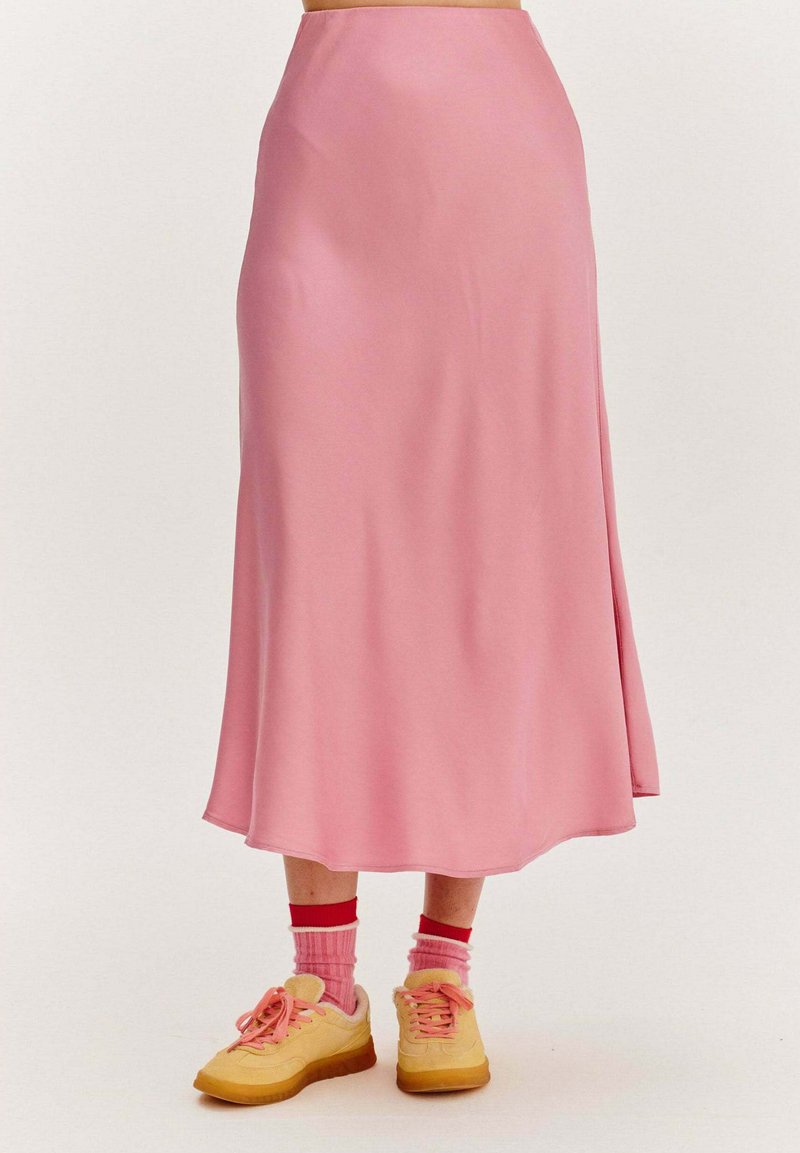 Ružová, splývavá midi sukňa vyrobená z hladkej tkaniny. Skombinovaná so žltými teniskami a ružovými ponožkami s červenými akcentmi. Jednoduchý dizajn, bez viditeľných vzorov.