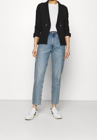 Osoba v černém blejzru, bílém tričku, světle modrých džínech s vysokým pasem a bílých teniskách s černými detaily, stojící na hladké podlaze.