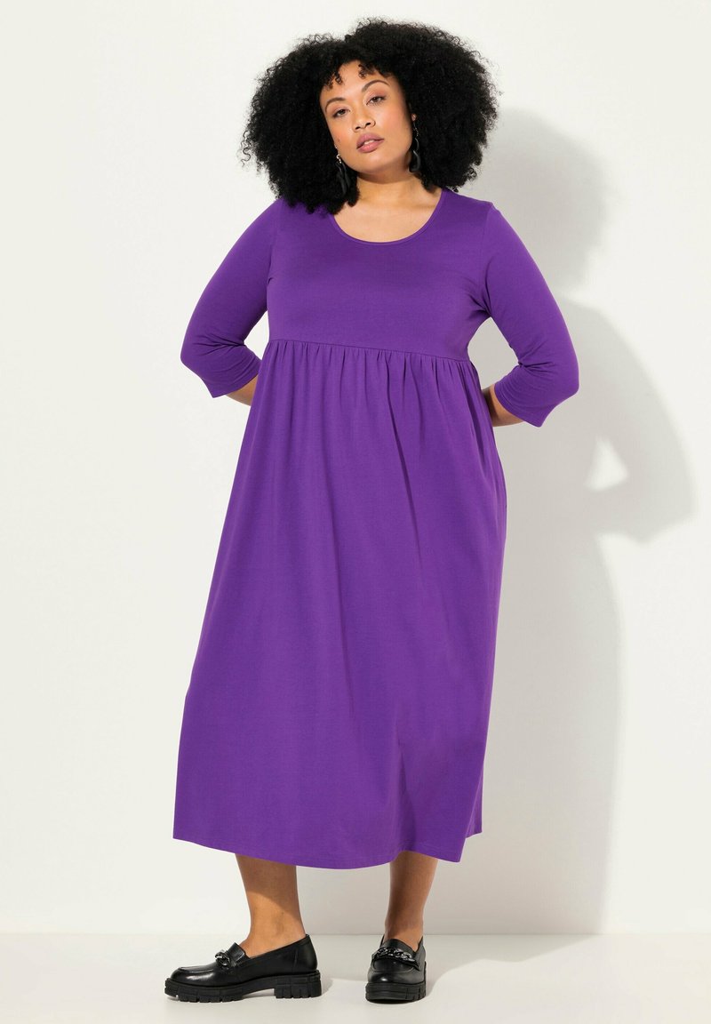 Robe violette, longueur genou, avec des manches trois-quarts et une taille froncée. Tissu lisse, associée à des chaussures noires à talons épais.