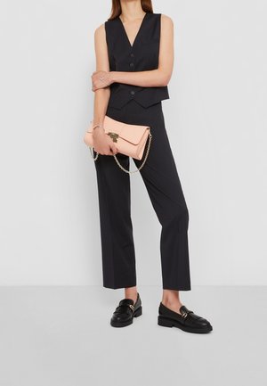 Person iført en ærmeløs sort vest og bukser, der holder en lys pink clutch med en guldkæde, iført sorte loafers.