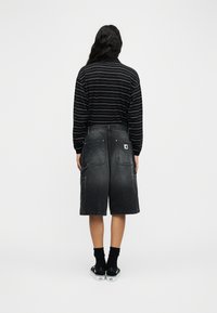 Άτομο με την πλάτη, φοράει μαύρο ριγέ μακρυμάνικο πουκάμισο, μαύρα τζιν σορτς μέχρι το γόνατο με μεγάλες τσέπες, μαύρες κάλτσες και μαύρα αθλητικά παπούτσια.