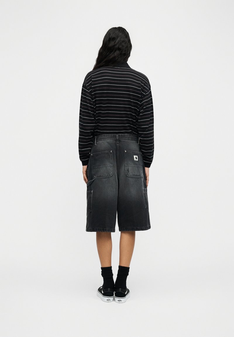 Person von hinten, trägt ein langärmeliges Hemd mit schwarzen Streifen, schwarze Denim-Shorts bis zum Knie mit großen Taschen, schwarze Socken und schwarze Turnschuhe.