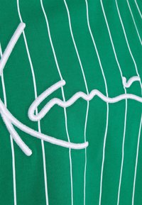 Zielona koszulka z krótkim rękawem w cienkie pionowe białe paski oraz wypukłym, białym, haftowanym napisem na przodzie. Materiał wydaje się gładki.