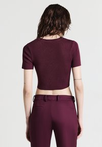 Persona con capelli castani bagnati di lunghezza spalla, che indossa una maglietta attillata a maniche corte di colore bordeaux e pantaloni a vita alta abbinati.