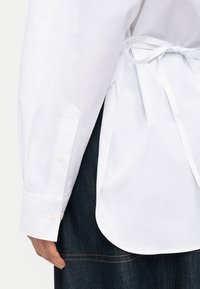 Κοντινό πλάνο ενός ατόμου που φοράει λευκό πουκάμισο με μακριά μανίκια με κουμπιά στα μανίκια και δεμένο πίσω, συνδυασμένο με σκούρο μπλε τζιν.