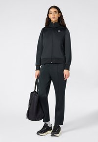 Czarna sportowa kurtka z pełnym zamkiem, ściągaczami na rękawach i logo. Połączona z pasującymi spodniami i czarnymi trampkami, trzymająca ciemną torbę.