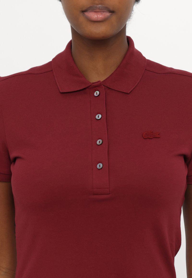 Polo burgundy en tissu texturé, avec un col, une patte de boutonnage avec des boutons contrastants et un logo brodé sur la poitrine.