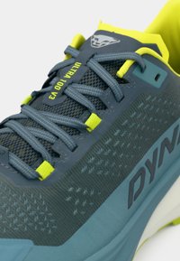 Μπλε και γκρι αθλητικό παπούτσι τρεξίματος με διχτυωτό επάνω μέρος, ανακλαστικές λεπτομέρειες και φωτεινές κίτρινες πινελιές. Διαθέτει έντονη σήμανση και υφές.