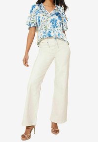 Žena v bílých širokých kalhotách, modro-bílé květinové blůze s krátkým rukávem a béžových lodičkách na vysokém podpatku, stojící s jednou nohou mírně vpřed.