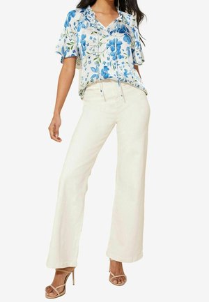 Femme portant un pantalon large blanc, un blouse à manches courtes bleu et blanc à fleurs, et des sandales à talons beiges, se tenant avec une jambe légèrement en avant.