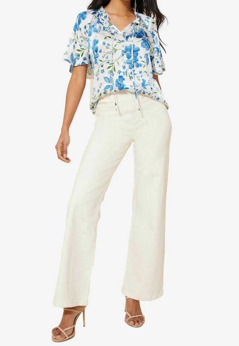 Žena v bílých širokých kalhotách, modro-bílé květinové blůze s krátkým rukávem a béžových lodičkách na vysokém podpatku, stojící s jednou nohou mírně vpřed.