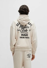 Felpa beige con una grande stampa nera sul retro, che presenta "Motor Club Est. '67 HUGO ENGINE PARTS." Tessuto morbido e vestibilità comoda.