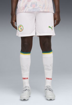 Person iført hvide Senegal fodboldshorts med hold- og Puma-logoer, hvide sokker med grønne og gule striber samt hvide fodboldsko.