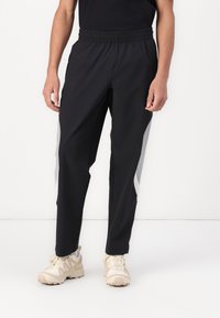 Sorte træningsbukser med elastisk talje, der har grå sidelogoer og en tapered design. Materialet virker let og glat.