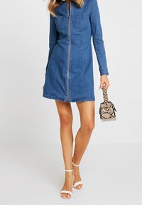 Jeansová košilová šaty s dlouhými rukávy, předním zipem, dvěma náprsními kapsami a přiléhavým střihem. Kombinováno s bílými sandály na podpatku a vzorovanou kabelkou.