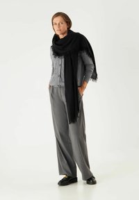 Cardigan gris avec des boutons, associé à un pantalon large gris. Une écharpe à franges noire drape les épaules. Des chaussures noires complètent la tenue.