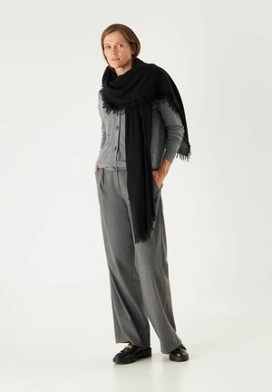 Grauer Cardigan mit Knöpfen, kombiniert mit grauen weitgeschnittenen Hosen. Ein schwarzer Fransen-Schal fällt über die Schultern. Schwarze Schuhe runden das Outfit ab.
