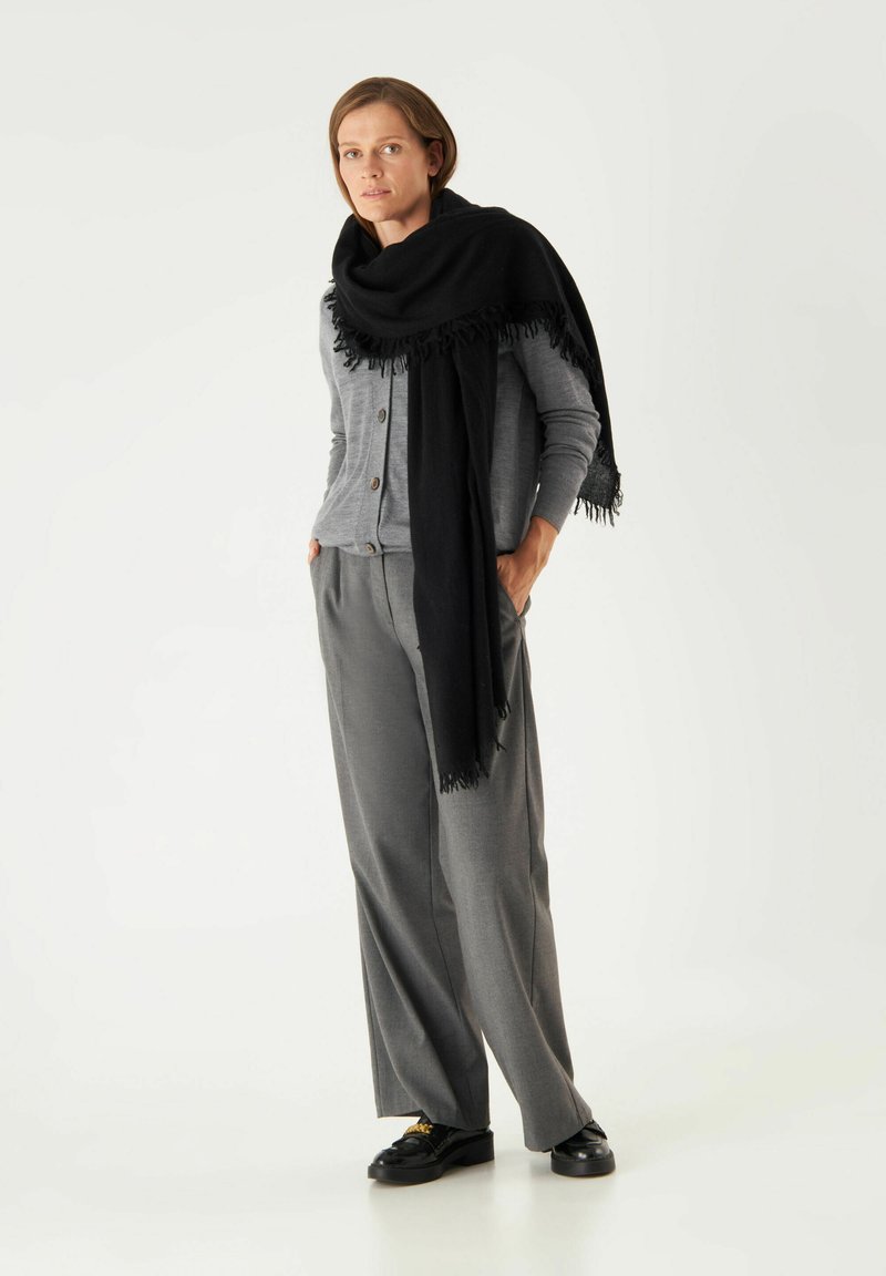 Cardigan grigio con bottoni, abbinato a pantaloni larghi grigi. Una sciarpa nera con frange pende sulle spalle. Scarpe nere completano l'outfit.