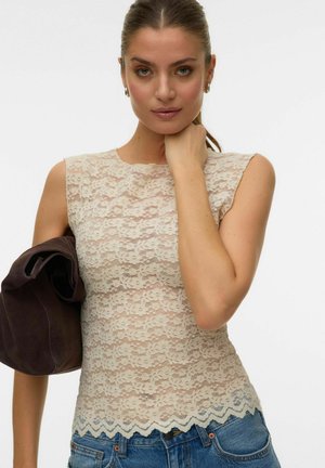 Vrouw met een paardenstaart, gekleed in een mouwloze beige kanten top en blauwe jeans, met een bruine jas over haar linkerschouder.