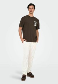 Brun bomulds T-shirt med en hvid grafik på venstre bryst, parret med hvide bukser og brune bådsko. Enkelt, afslappet design.