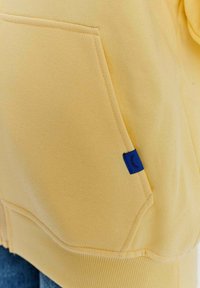 Žltý sweatshirt s veľkou prednou kapsou a modrou textilnou nálepkou. Materiál je mäkký a hladký s ribbovaným lemom.