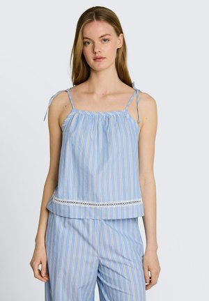 Młoda kobieta ubrana w niebiesko-biały pionowo paskowany bezrękawnik z wiązanymi ramiączkami i pasujące spodnie na jednolitym tle.