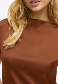 Brahns, īsas piedurknes blūze ar gludu, matētu struktūru. Apaļa kakla izgriezuma forma un vienkāršs dizains, minimālas detaļas redzamas.