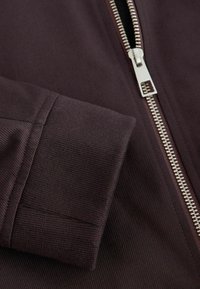 Gros plan d'une manche de veste en tissu côtelé marron foncé avec une fermeture éclair métallique argentée à curseur rectangulaire.