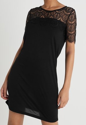 Femme portant une courte robe noire avec de la dentelle couvrant les épaules, le haut de la poitrine et les manches, debout devant un fond uni.